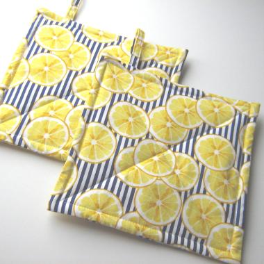 2 overlapping squares of padded fabric with thin vertical blue and white stripes partially covered by randomly placed images of yellow lemon slices. Hanging tabs on the upper edge.